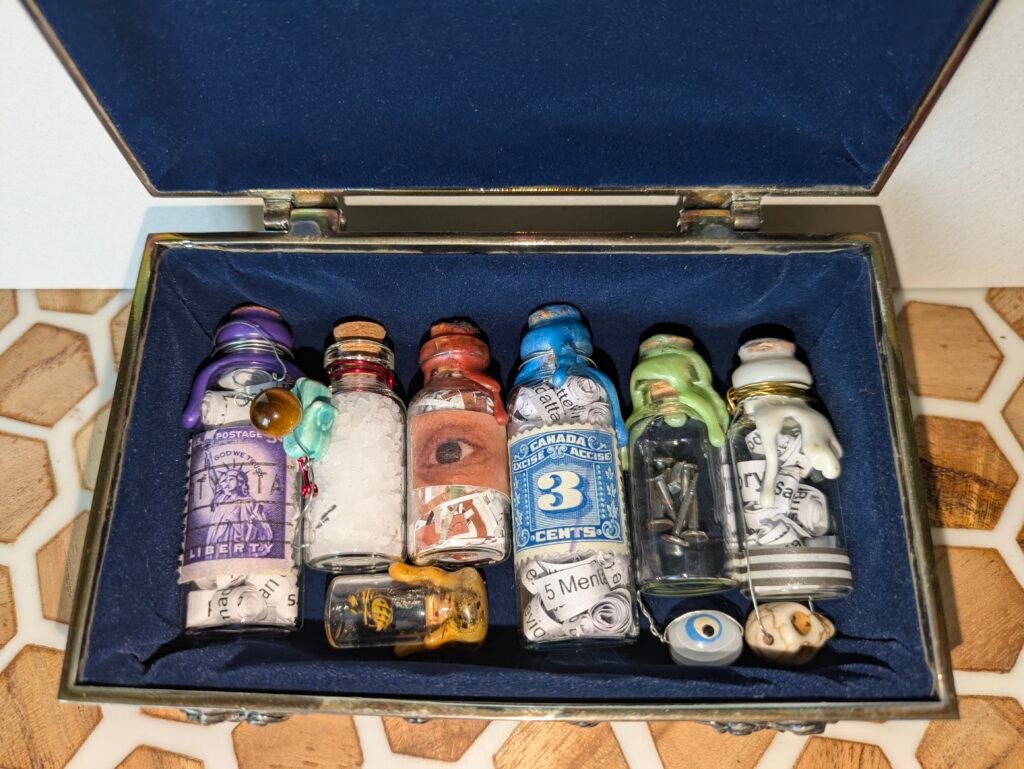 Four two inch and four one inch mini bottles are lined up in a velvet blue box. Each is corked and sealed with wax, decorated with stamps, images and beads. The images include skulls, eyes, nails and the statue of liberty. Inside the bottles are rolled paper, salt and a wasp.