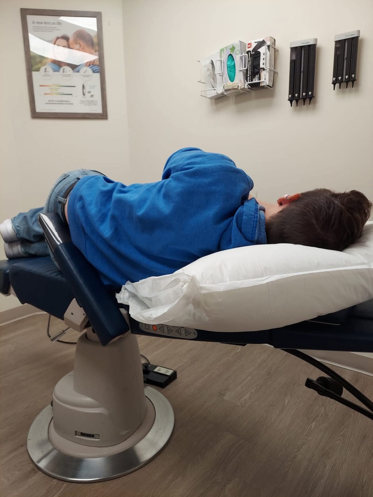 A person lays on a reclined medical chair with a pillow under their head. They are in a medical office and there are disposable gloves, ear canal tips and other supplies.