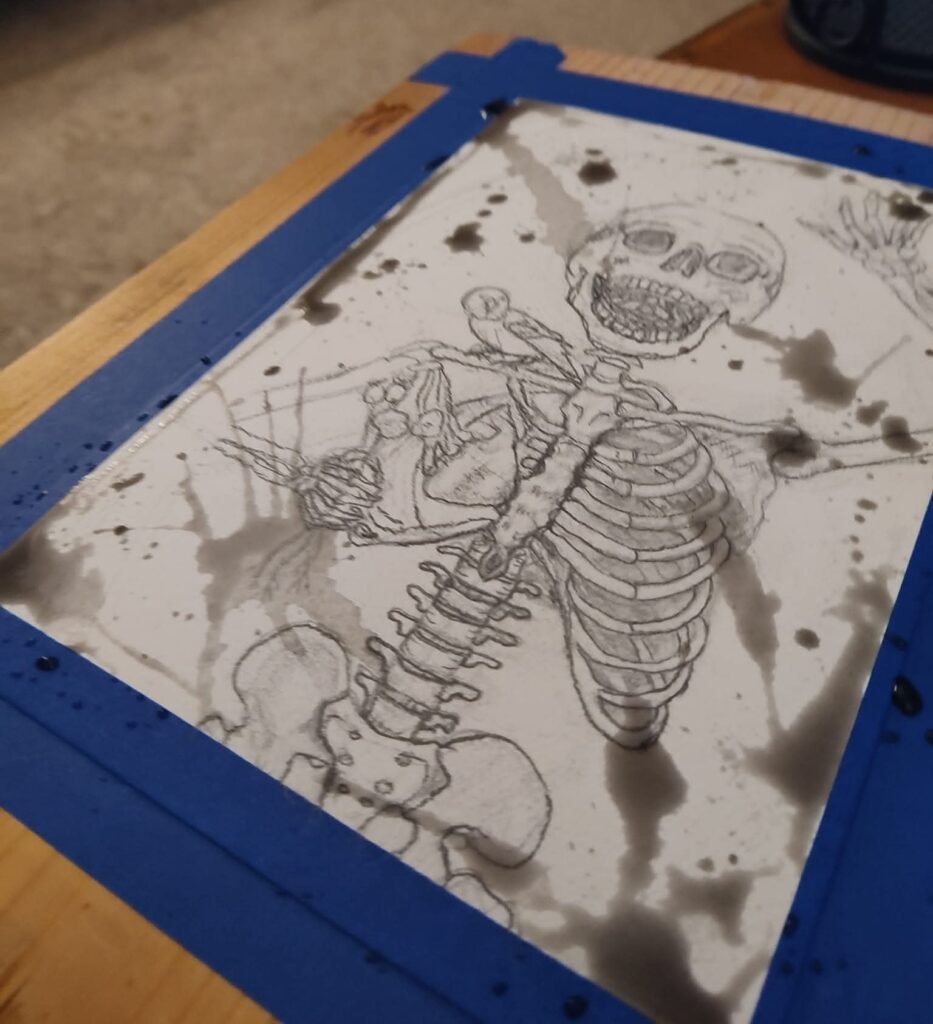 A skeleton with a bird on it's shoulder is drawn in pencil and taped with blue tape to a board. There are splashes of wet, black ink all over the page.