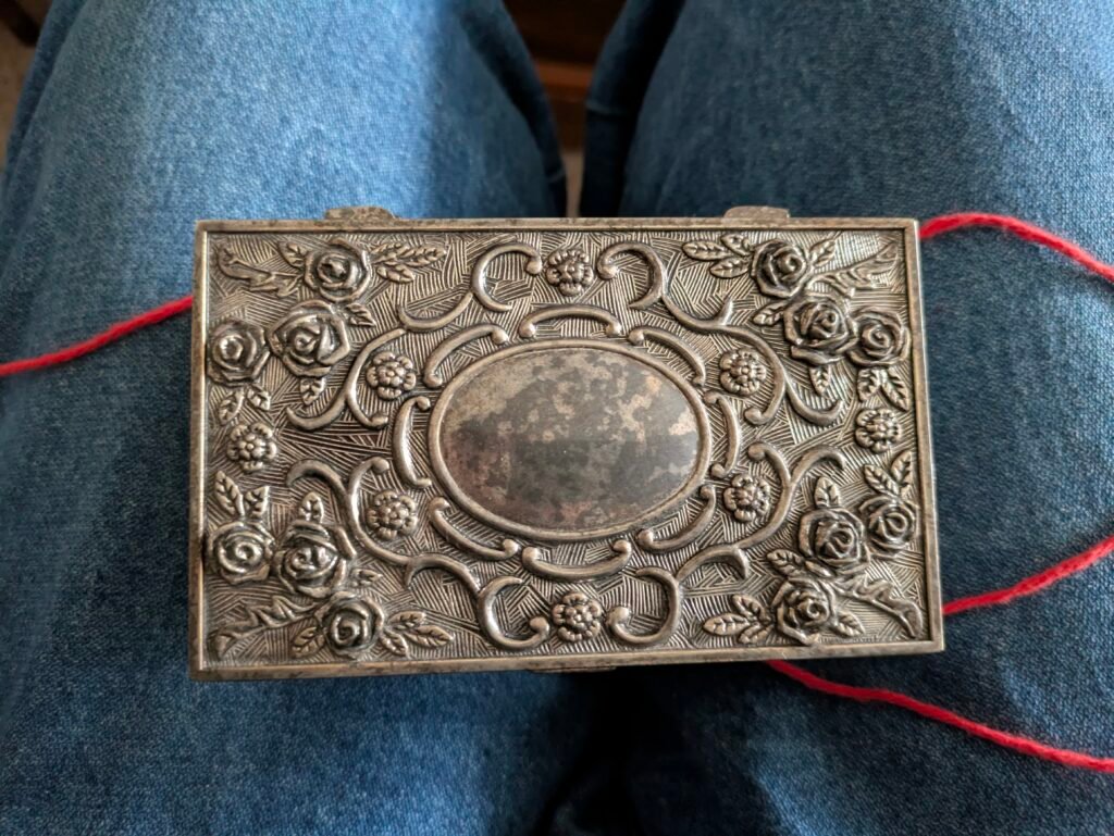 Silver plated jewelry box with roses carved around the box. It sits on a lap wearing blue jeans and a red string has been untied & sits under the closed box