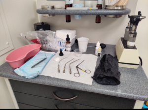 A counter is covered in medical bottles and instruments. There are three hook like metal straws, tweezers and eye droppers. There are black gloves and gauze too.